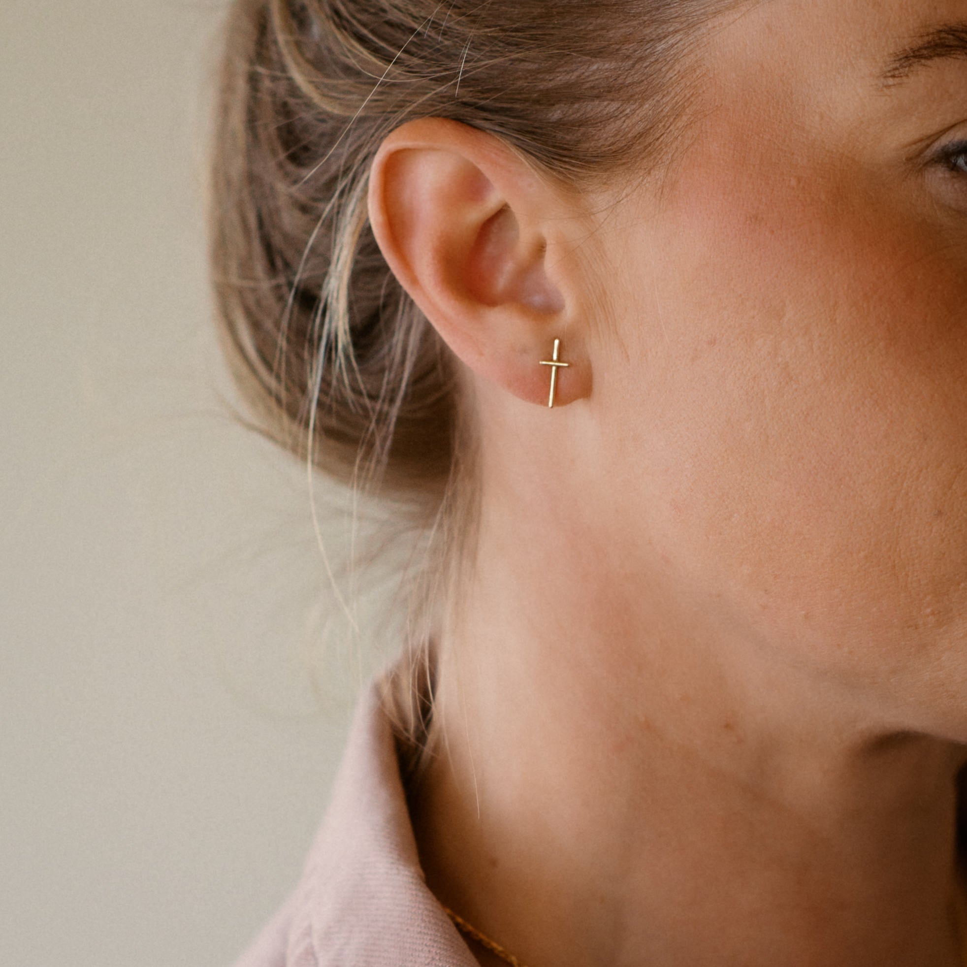 Close-up of a person wearing a 18K gold cross earring with a neutral background