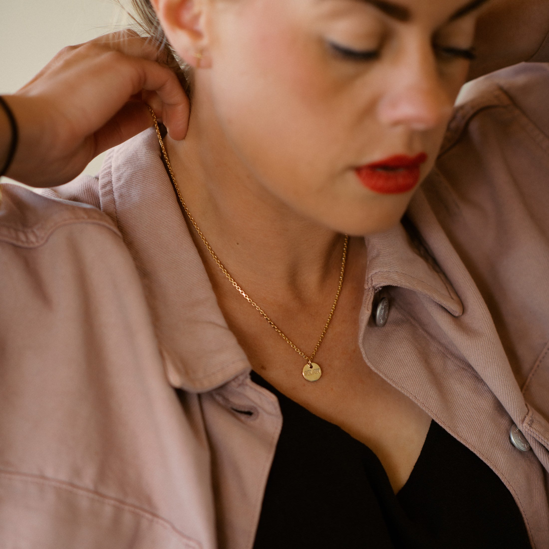 Woman wearing a gold necklace with a small pendant, adjusting her jacket.