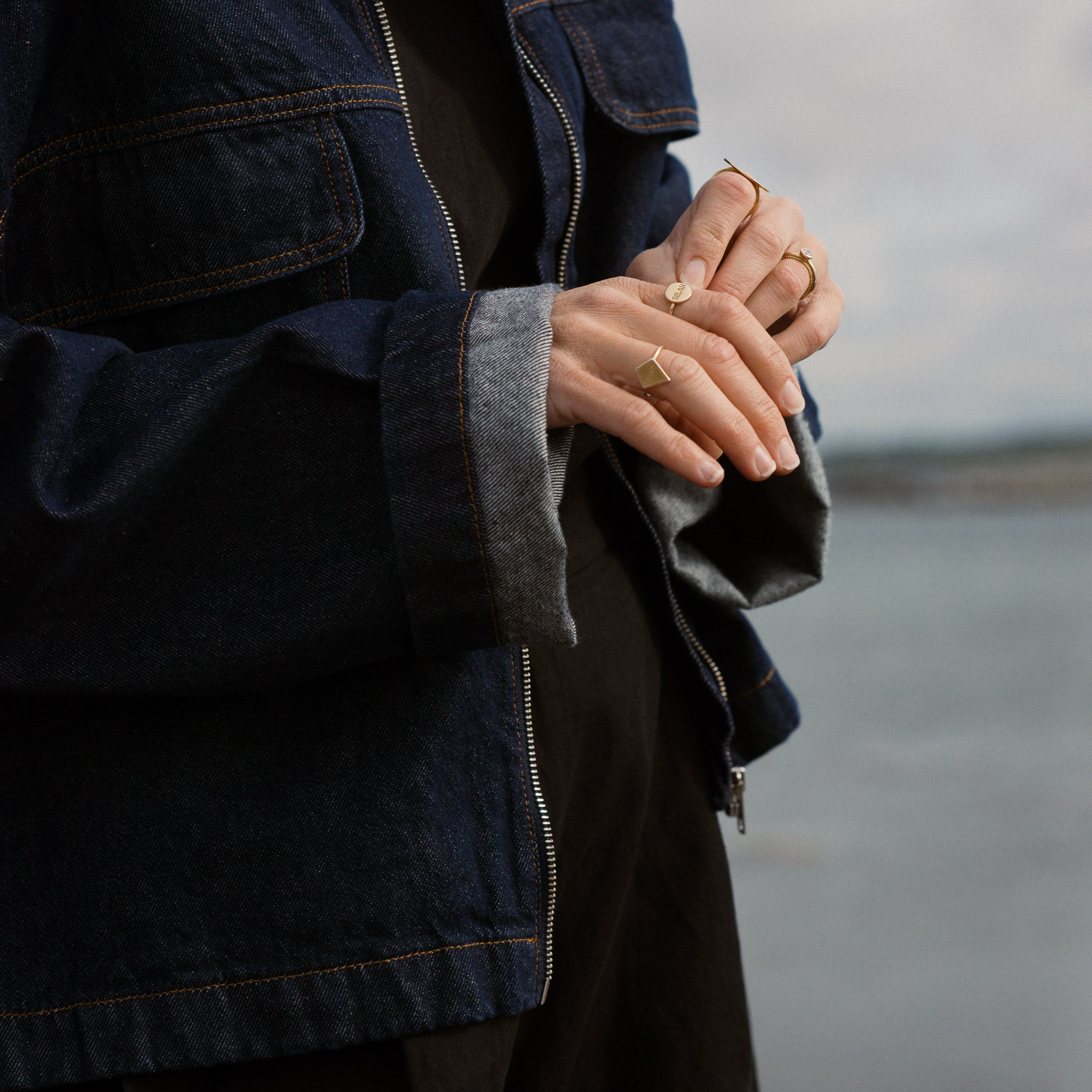 Person wearing a denim jacket with visible jewelry, standing against a blurred background.