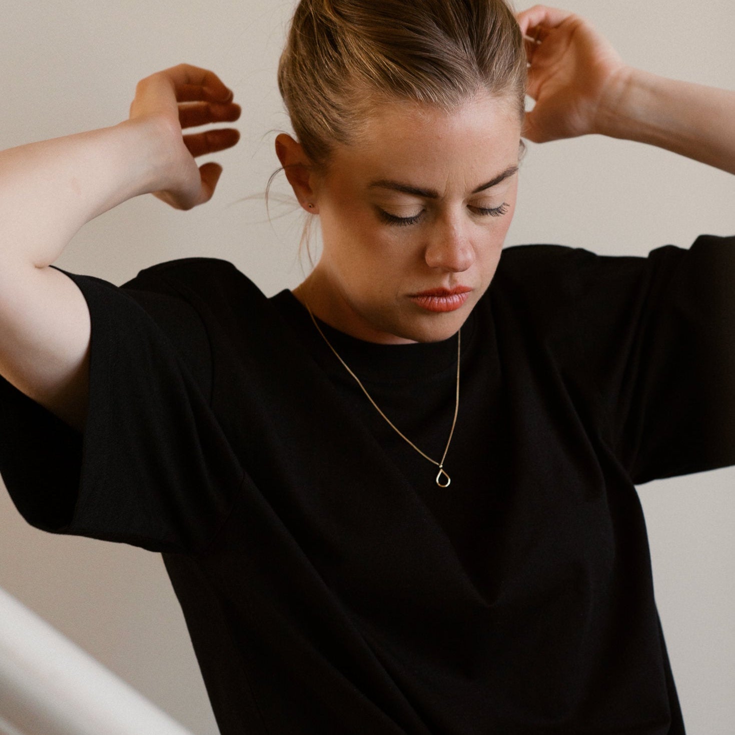 Woman adjusting her hair in a black shirt against a neutral background, 18K tears of hope gold necklace.