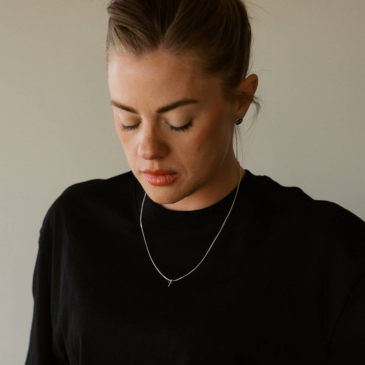 A woman wearing a black shirt and a necklace against a neutral background, with a small white gold cross necklace